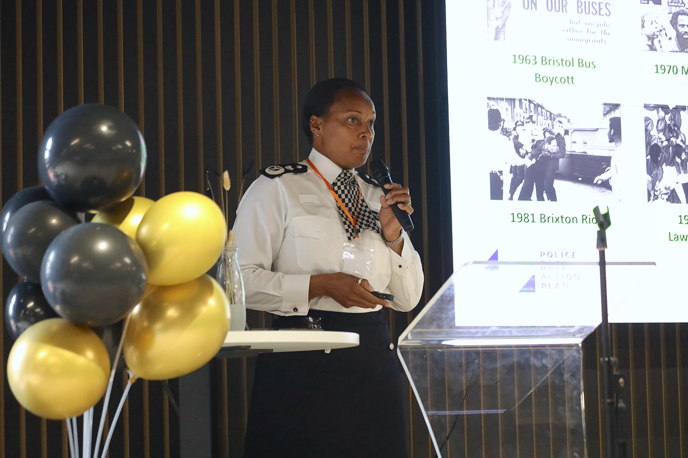 A female people officer is stood on a stage with a microphone in her hand. There is a presentation on a screen behind her.