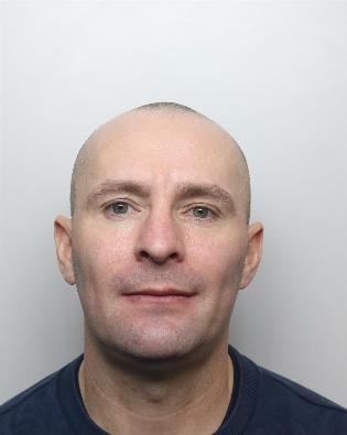 A bald men seen smirking looking towards the camera, wearing a black t-shirt and stood against a grey background.