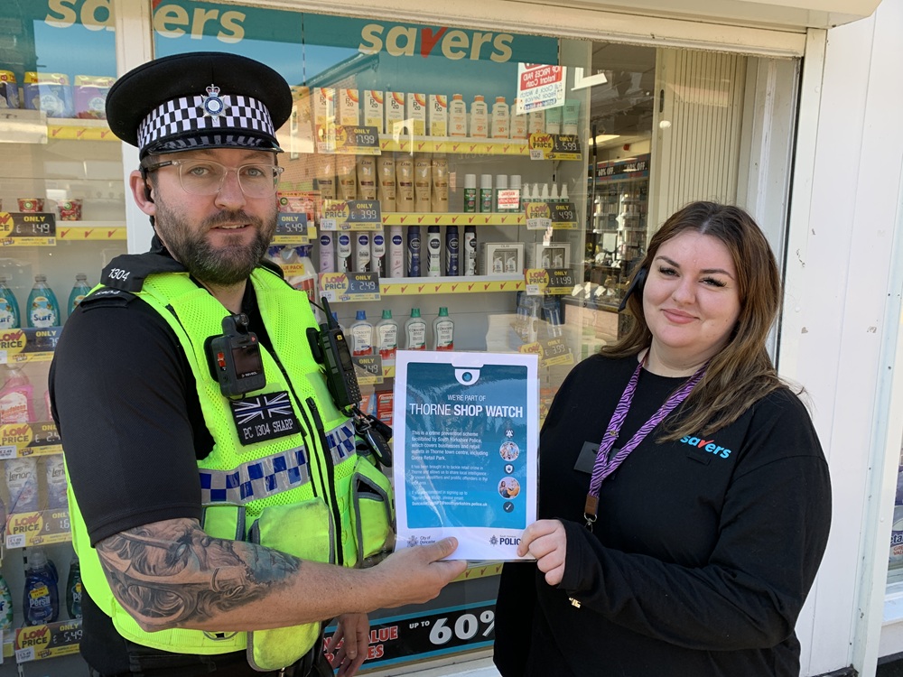 A police officer shakes a shop worker's hand as they hold up a piece of paper promoting Thorne Shop Watch