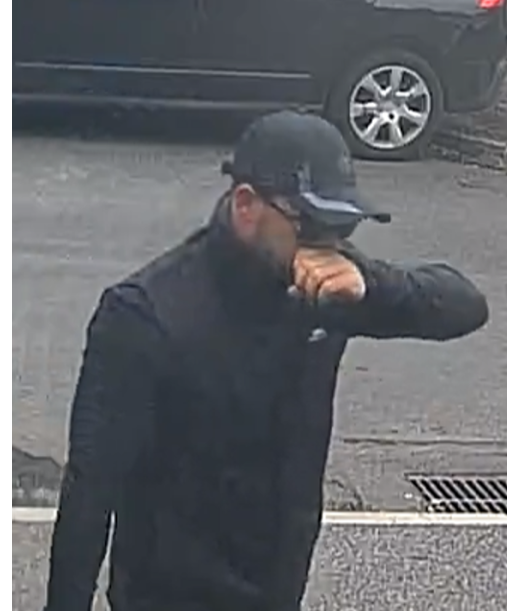 A man with short dark brown hair and in his 30s. He is wearing a black tracksuit, black cap, and dark sunglasses.  
