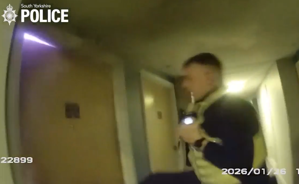 A police officer is shown in a dimly lit corridor holding a torch while standing beside a closed door. The South Yorkshire Police logo appears in the top left corner, and body‑worn video timestamps are visible at the bottom of the image.