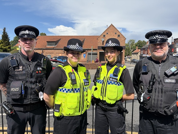 Three police officers stand outside a brick building with parked cars behind them