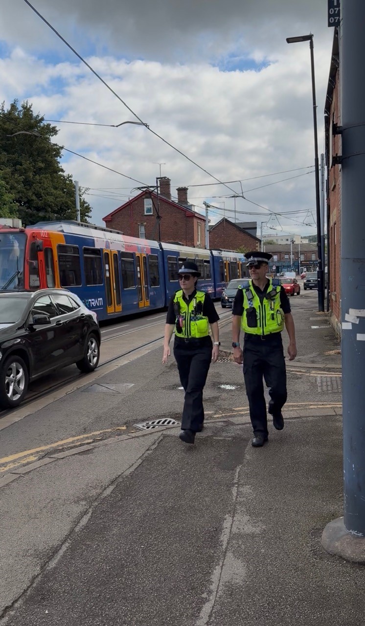 Two police officers in high visibility vests walk on a street with a tram in the background
