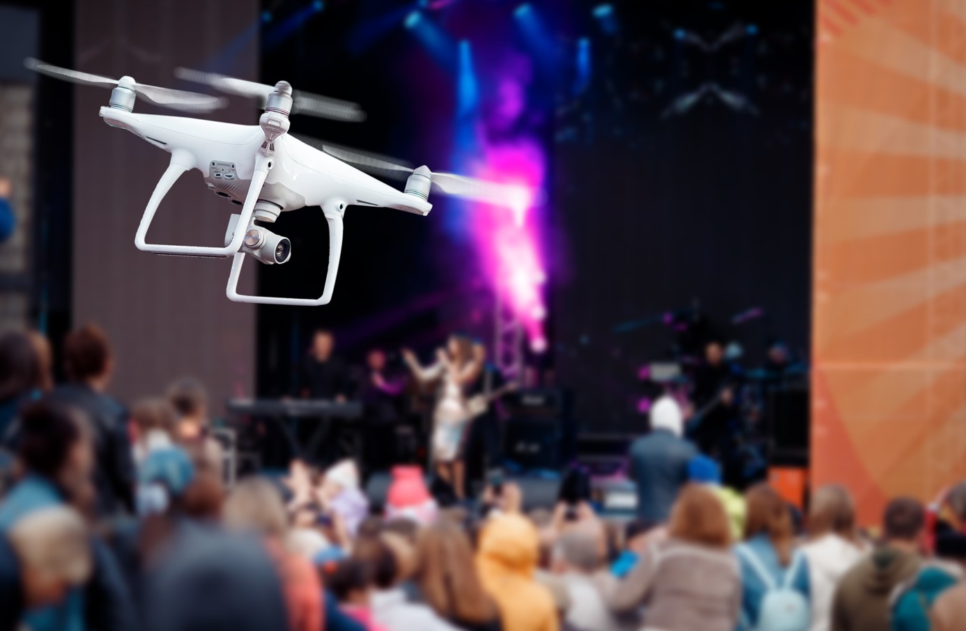 A white drone flies over a crowded music festival while someone performs in the background