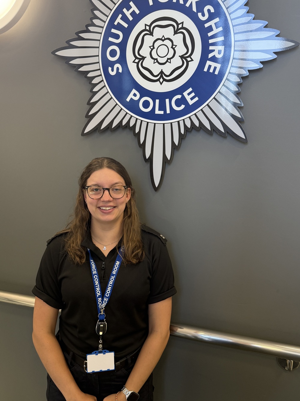 A smiling woman in a South Yorkshire Police uniform standing in front of the force's emblem on a grey wall.