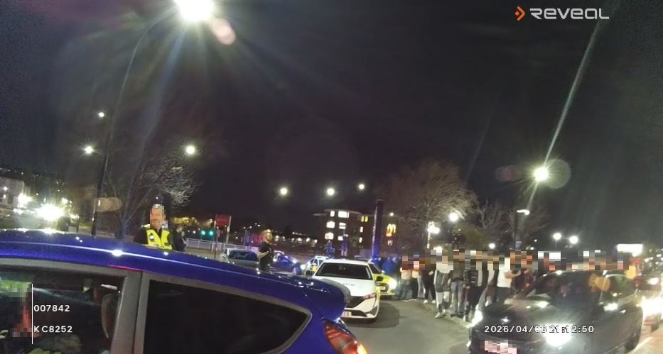 A screenshot of body worn video of people standing and cars gathering at a car meet