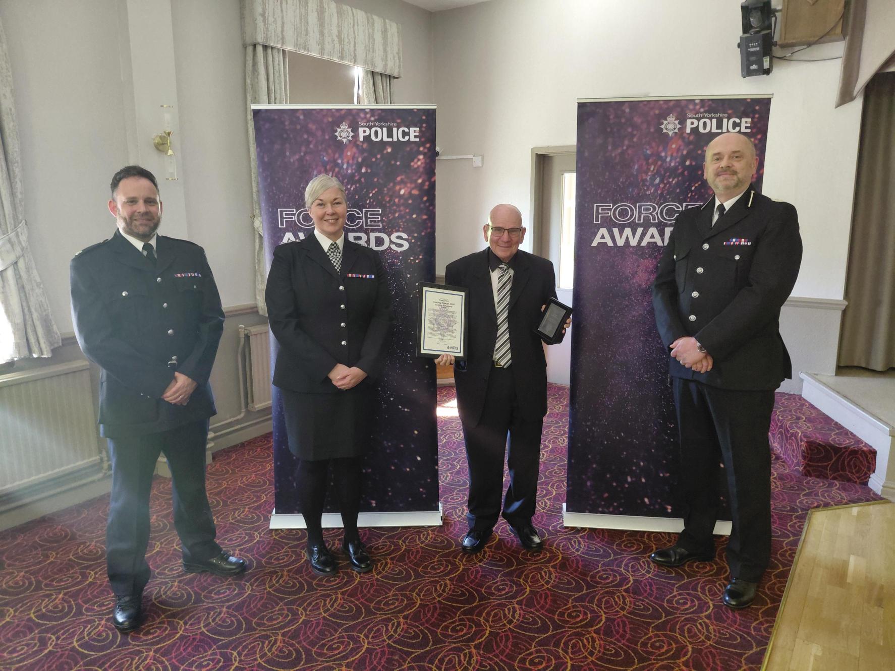 Three males and a female stand together wearing all black, with the male in the middle holding a certificate and an award