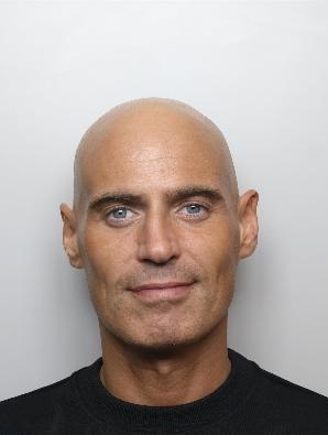 Gavin Bennett is a white man with a bald head and wearing a black t-shirt. He is standing in front of a white background for a custody picture