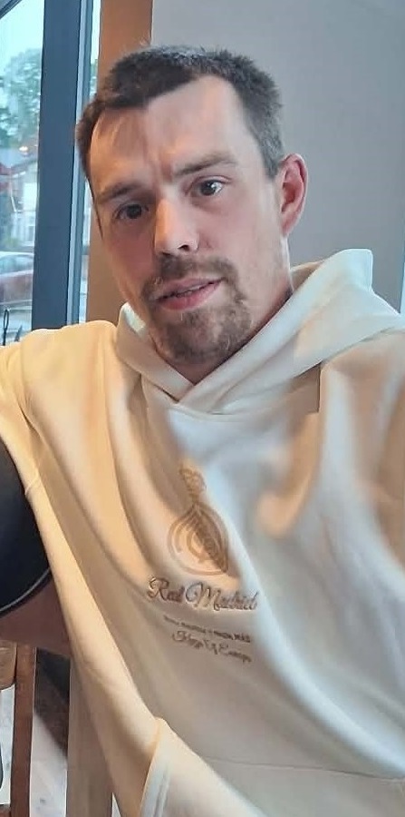 A man with short dark hair and a beard wearing a cream-coloured Real Madrid hoodie, looking at the camera