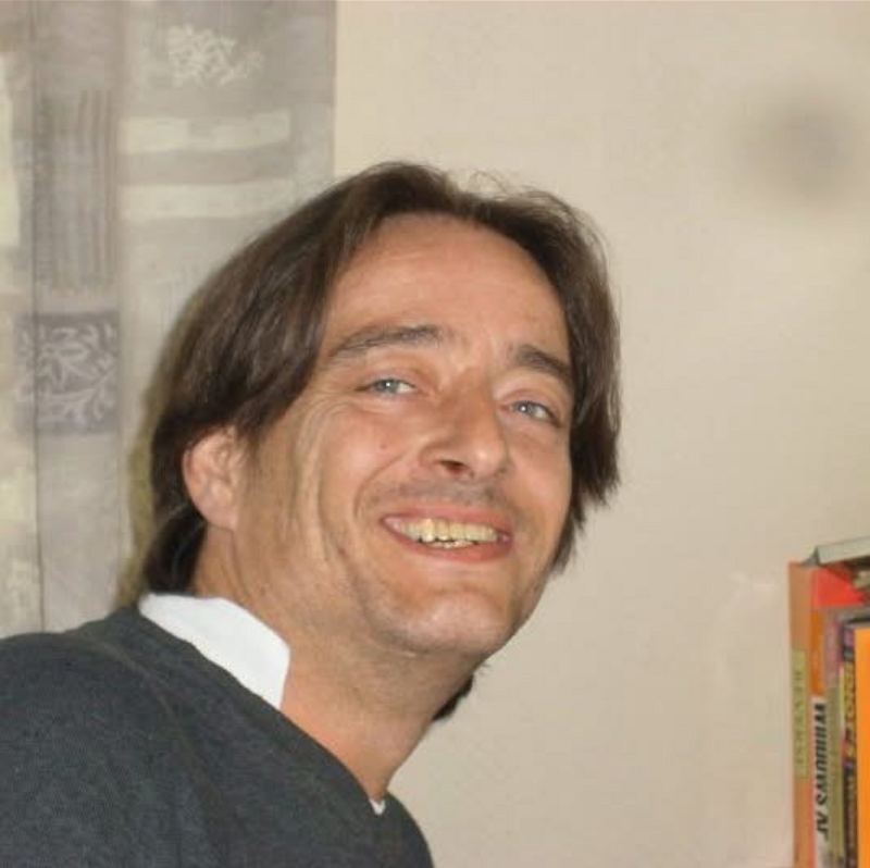 A smiling middle-aged man with shoulder-length dark hair wearing a grey jumper and white shirt