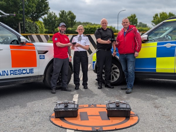 Four people, including police and mountain rescue personnel, hold drone controllers next to emergency vehicles and drones.