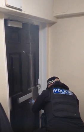 A police officer wearing a helmet uses a chainsaw to cut vertically through a door