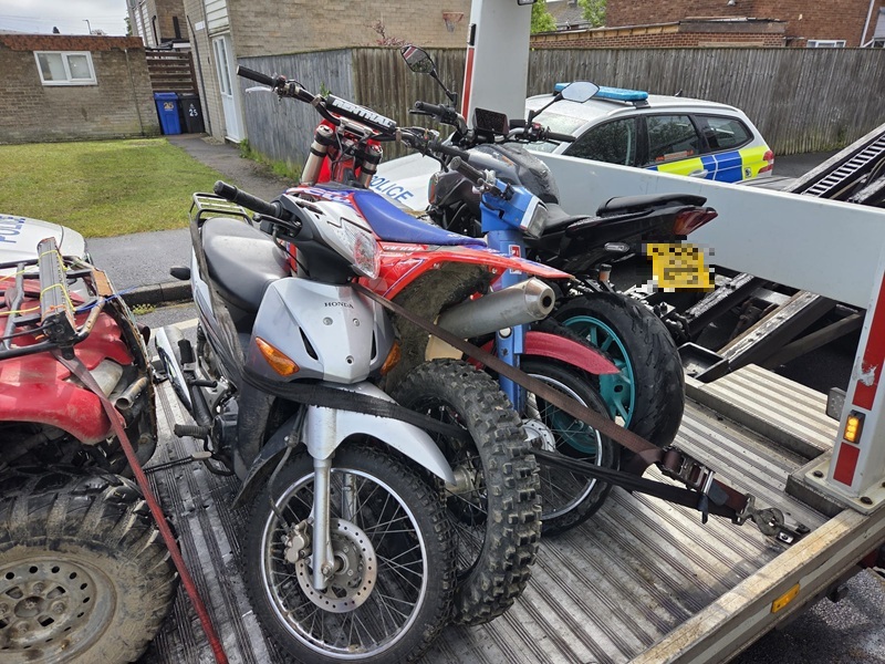 Off-road bikes and a quad bike are strapped onto a flatbed recovery truck, with a police car parked nearby on a residential street