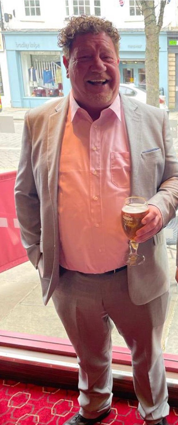 Jason Gaskin smiles and poses for a picture while holding a beer. He was a white man in his 50s, of a large build, with curly ginger hair