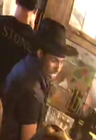 A man wearing a black trilby hat and a dark t-shirt in a bar