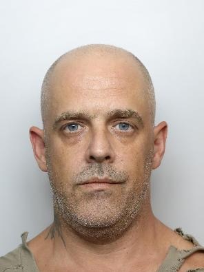 Custody image of a white middle-aged man with very short/nearly bald hair, grey short stubble and a t-shirt ripped at the top